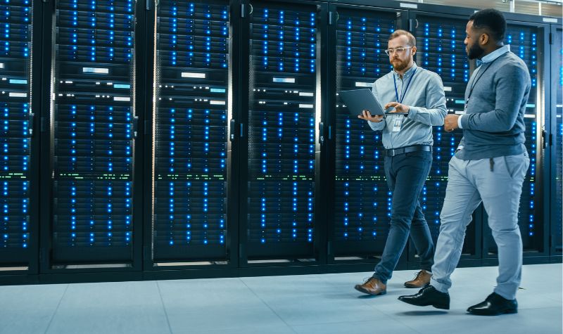 IT Technician with a Laptop Computer and Black Male Engineer Colleague are Talking in Data Center while Walking Next to Server Racks